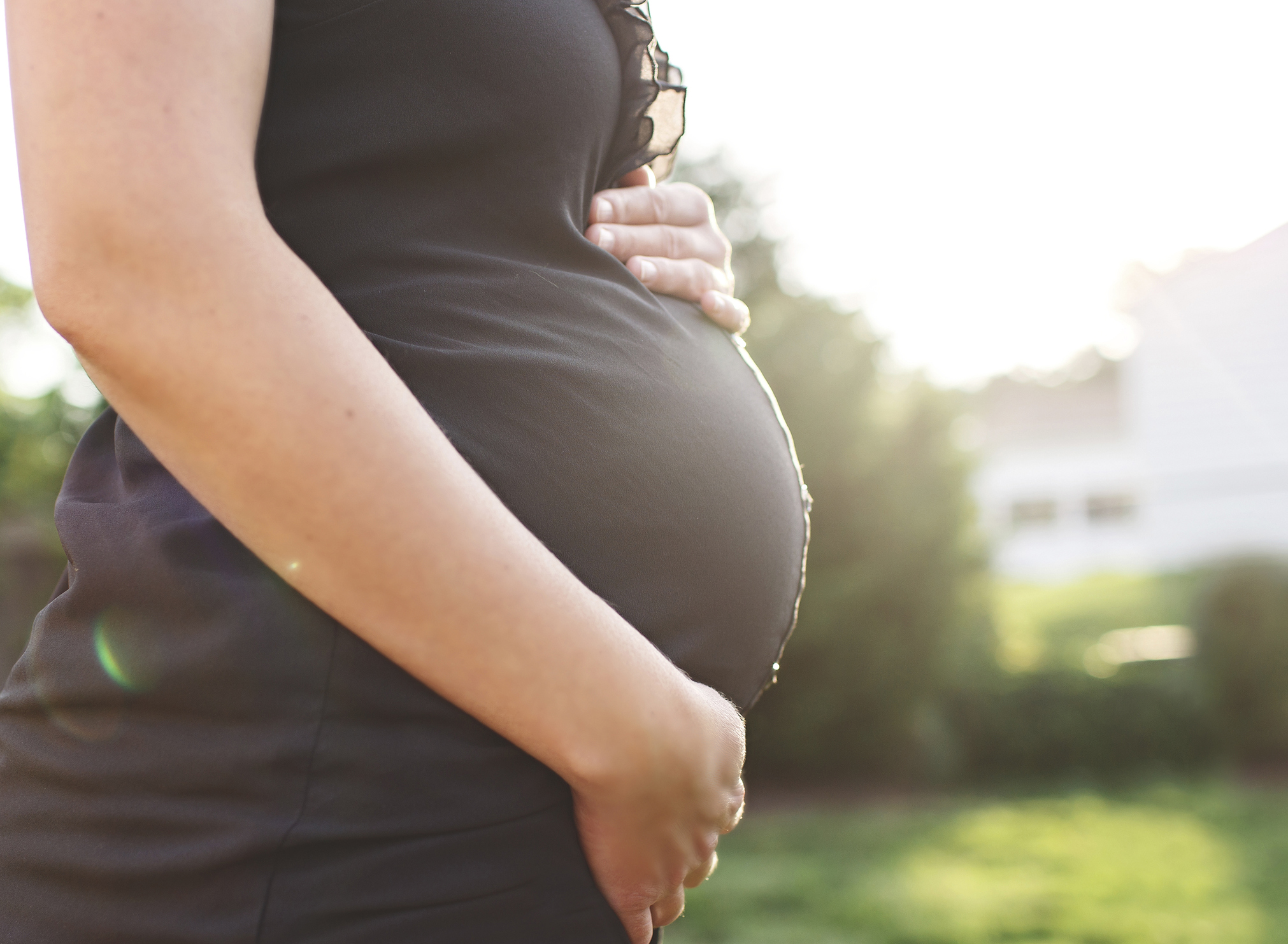 Side view of pregnant woman standing outdoors, one hand on top of her pregnant belly and one hand underneath the bump.
