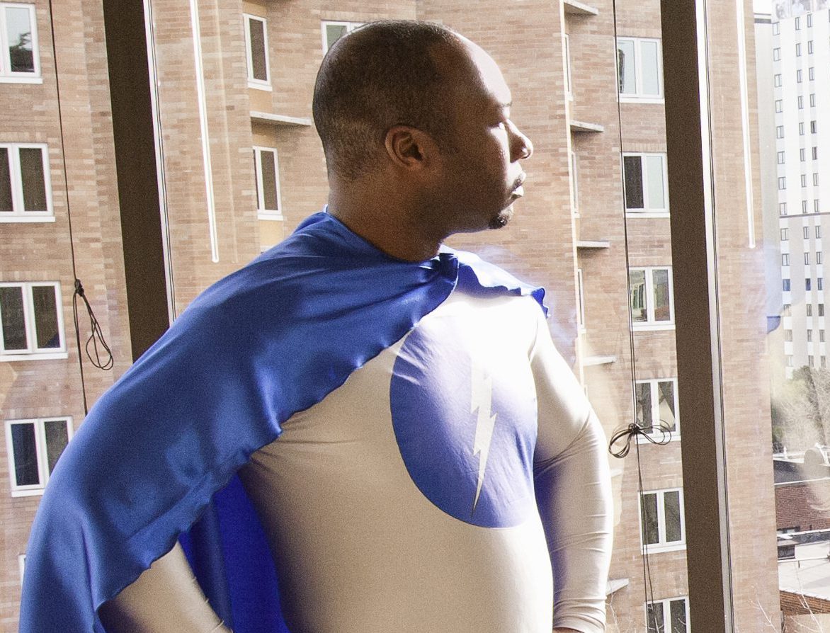 A black businessman office super hero stands at an office window and ponders his next business move.